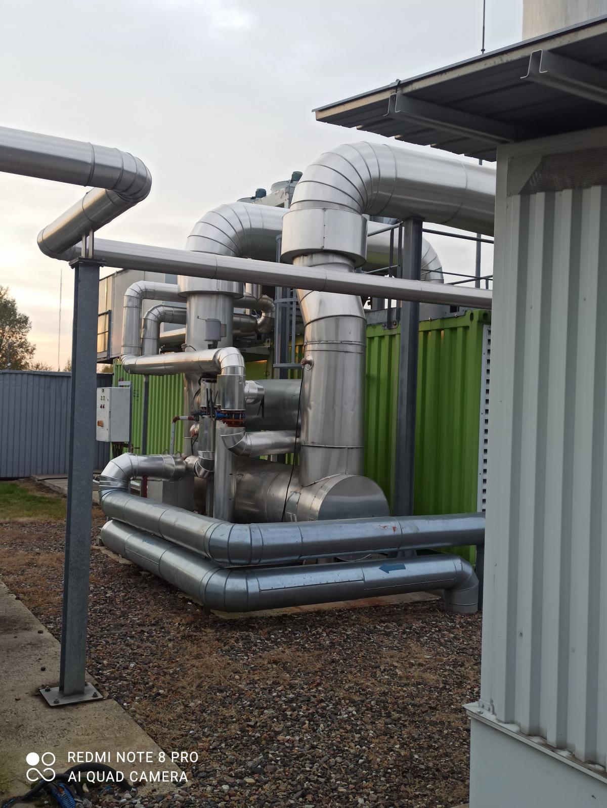 Złożona instalacja przemysłowa z rurami, zbiornikami i kontenerem. Elementy metalowe w otoczeniu żwiru i budynk&oacute;w.